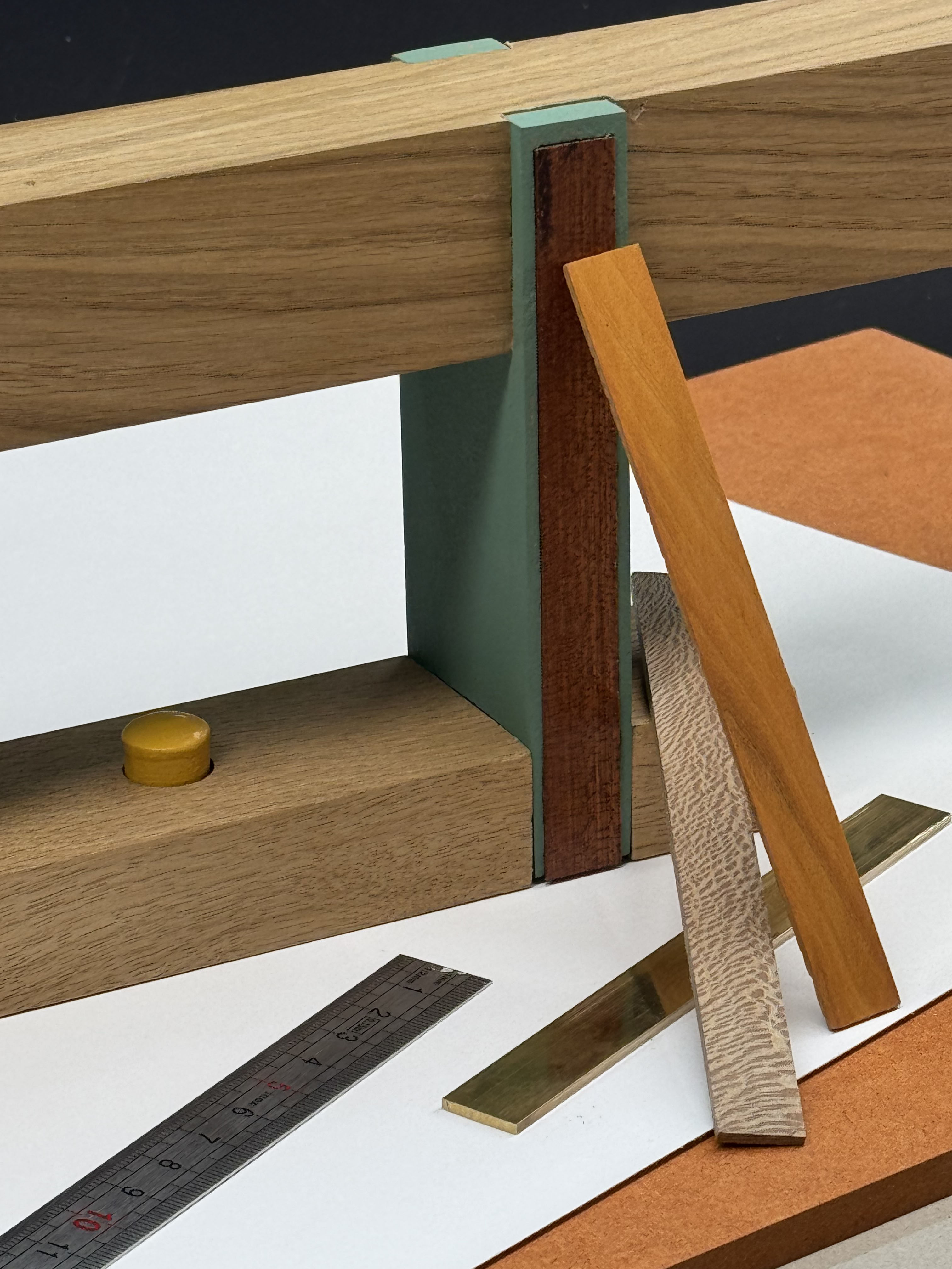 Close-up of a woodworking setup featuring a wooden carpenter's plane with green and brown detailing, three narrow wood samples leaning against it, and a metal ruler on a white surface with a brown background.
