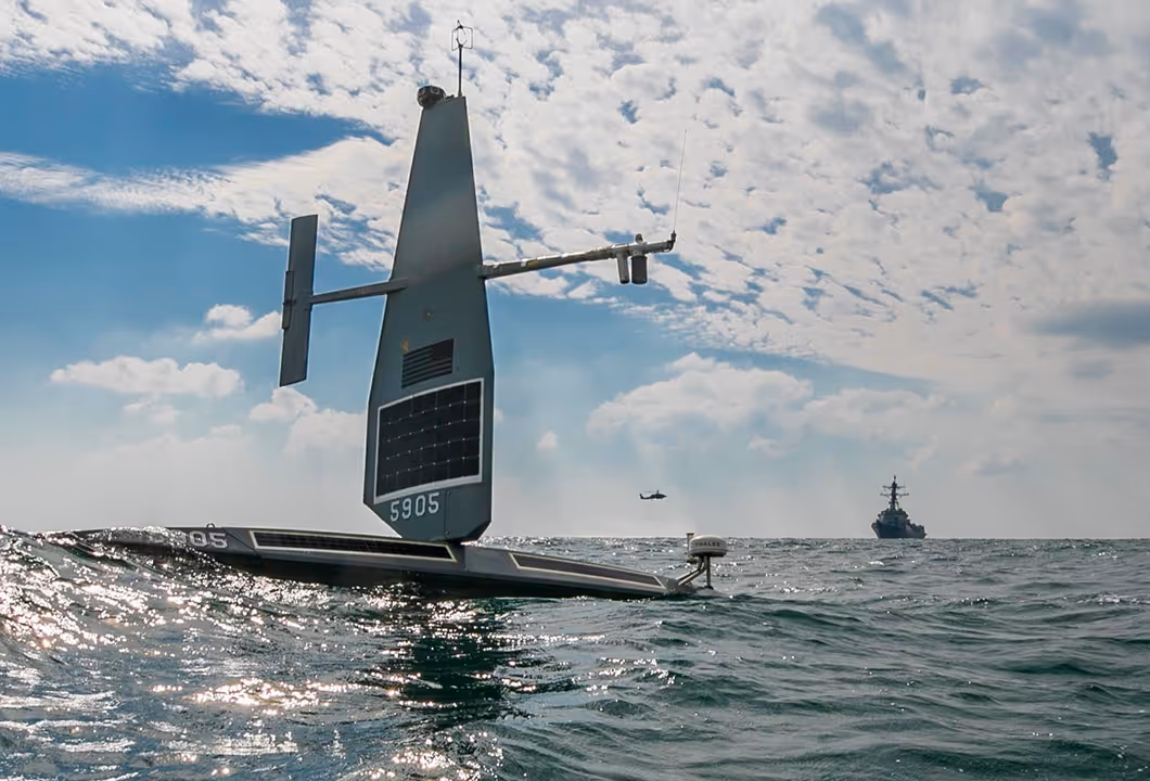 Autonomous unmanned surface vehicle with a vertical sail and solar panels floating on the ocean, with a helicopter and a naval ship in the distant background.