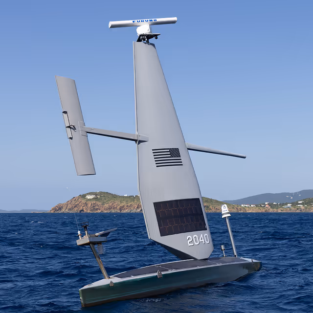 Autonomous Saildrone with a large vertical sail marked 2040 and an American flag, sailing on blue ocean water near a rocky island under a clear sky.