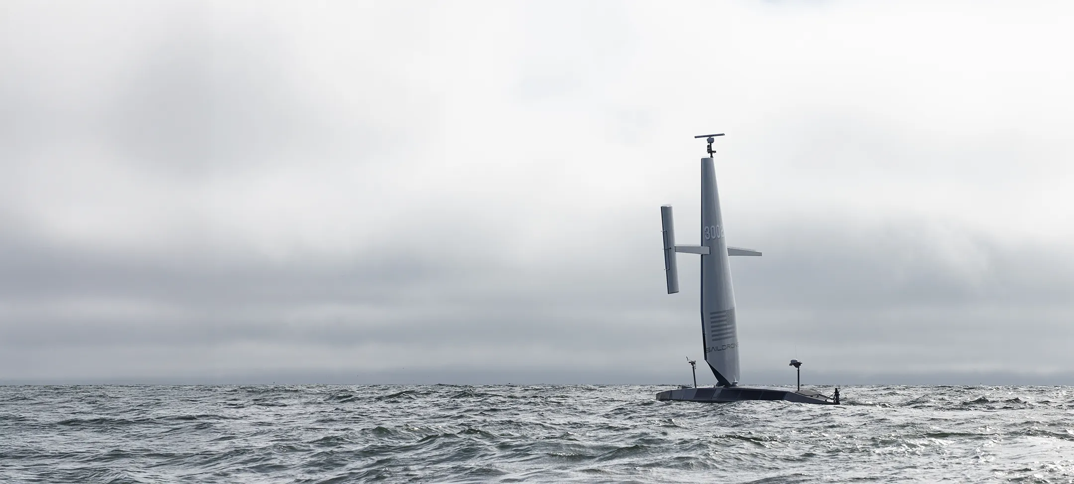 Unmanned Saildrone with tall vertical sail floating on a choppy ocean under cloudy sky.