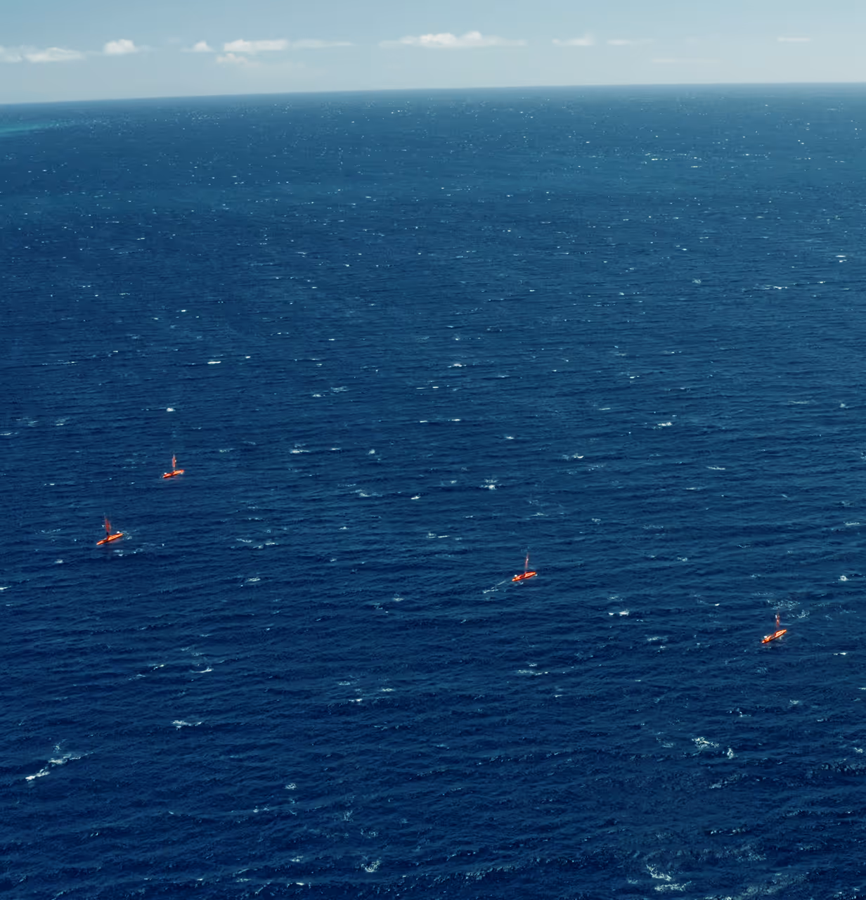 Four orange Saildrones sailing across a vast, deep blue ocean under a partly cloudy sky.