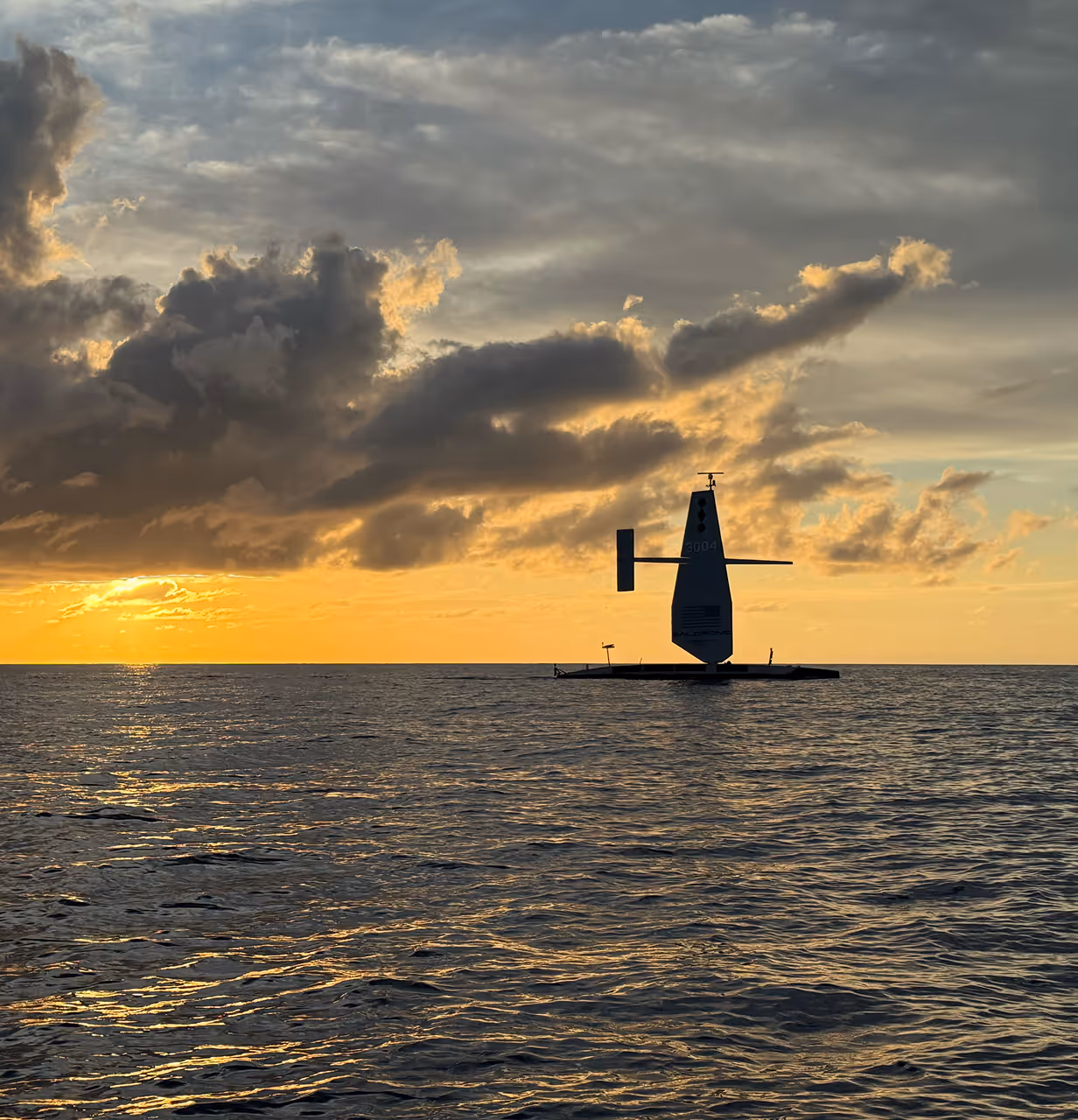 Silhouette of an Saildrone on calm seas at sunset under dramatic cloud-filled sky.