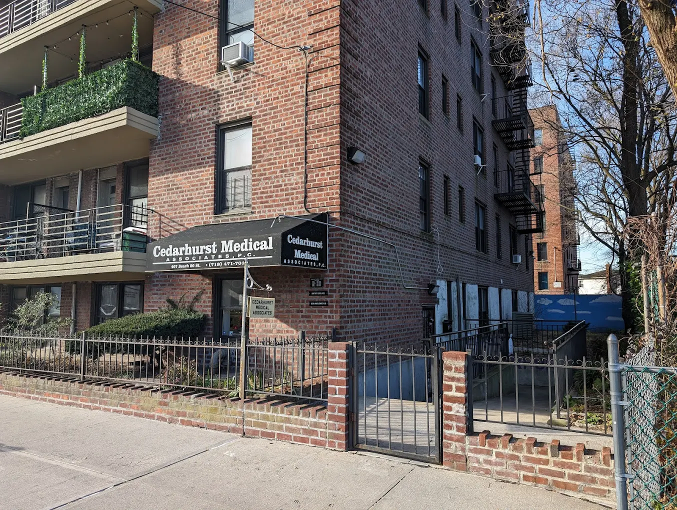 Street-level entrance to Cedarhurst Medical Associates office located on the ground floor of a large brick building with a black awning over the door displaying the phone number 718-471-7010.