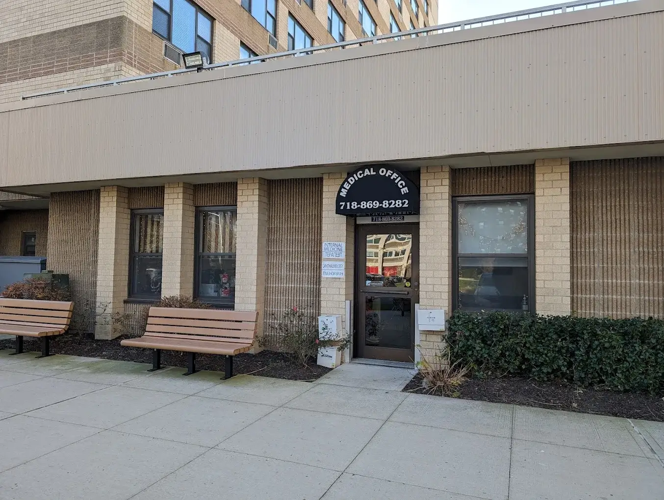 The front entrance to Cedarhurst Medical Associates office with a light brick facade, two brown benches, and a sign above the door displaying the phone number 718-869-8282.