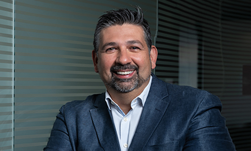 Smiling man with dark hair and beard wearing a blue blazer and white shirt, standing with arms crossed in front of a glass wall with horizontal stripes.