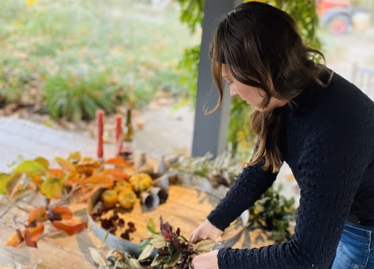 ChatGPT said:  Woman arranging an autumn wreath with colorful leaves and dried foliage on an outdoor wooden table.