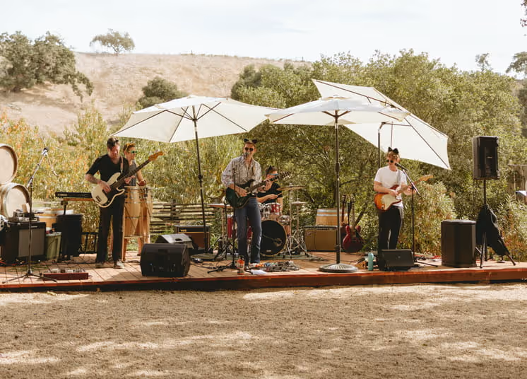 Live band performing on outdoor wooden stage with white umbrellas at rustic vineyard venue surrounded by oak trees