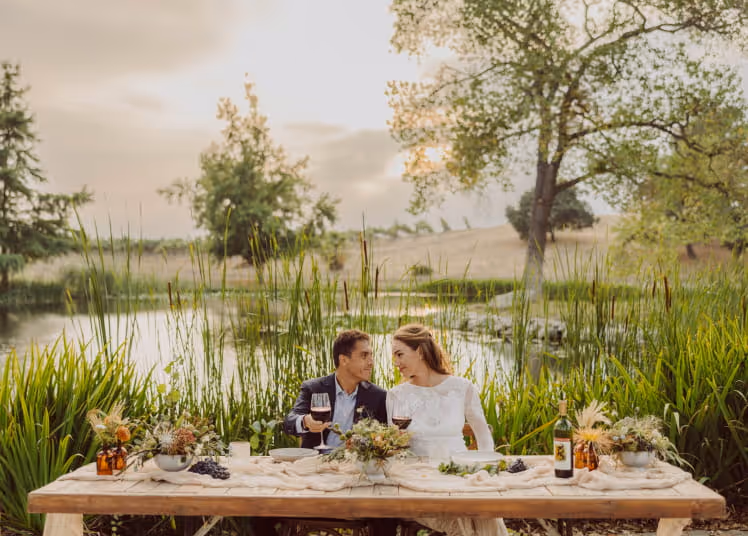 Couple sharing red wine at a rustic outdoor table decorated with flowers beside a scenic pond at golden hour