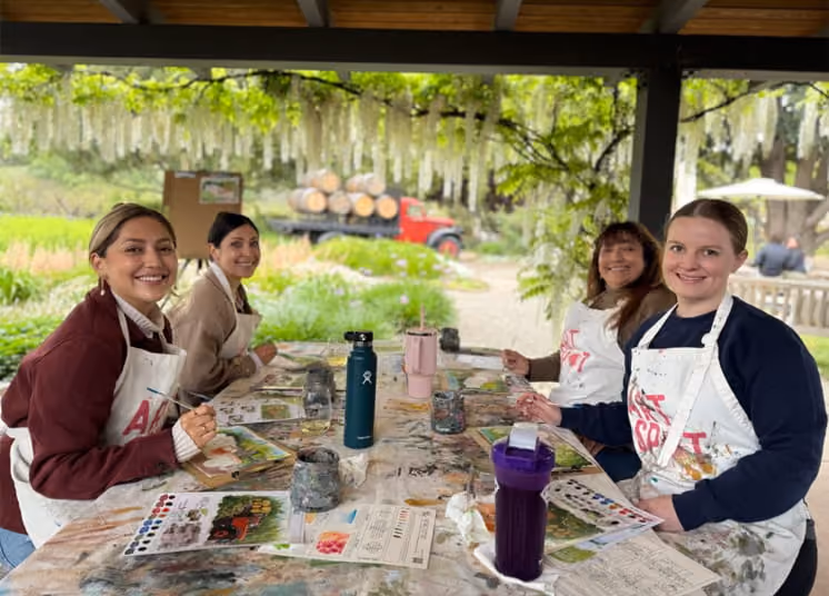 Four smiling women wearing aprons participate in an outdoor watercolor painting class at a vineyard with wisteria blooming