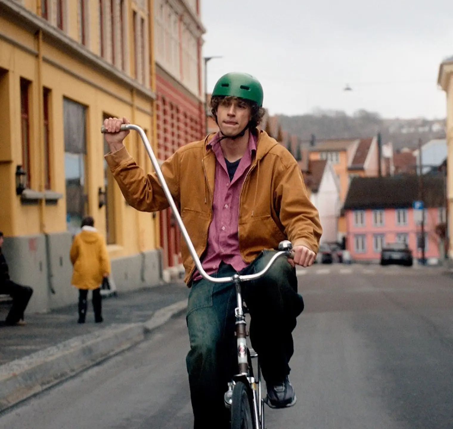 En ung man med grön hjälm cyklar på en gata i stan på en cykel med högt styre.