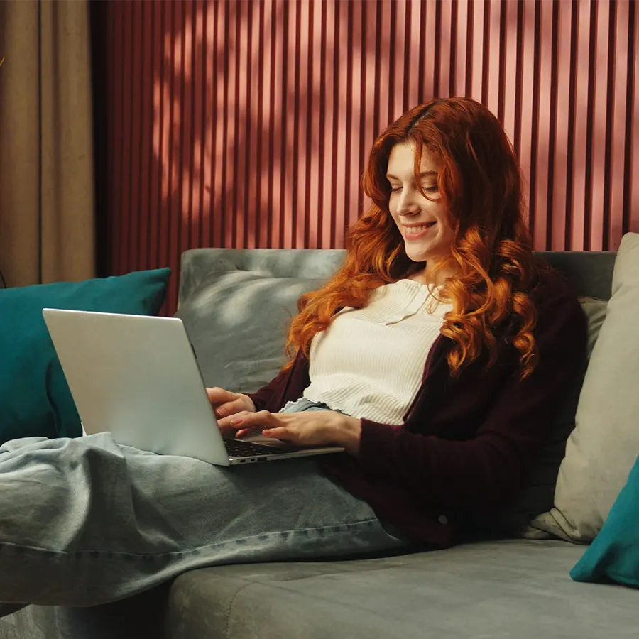 Woman with red hair using a laptop on a cozy sofa in a stylish room.