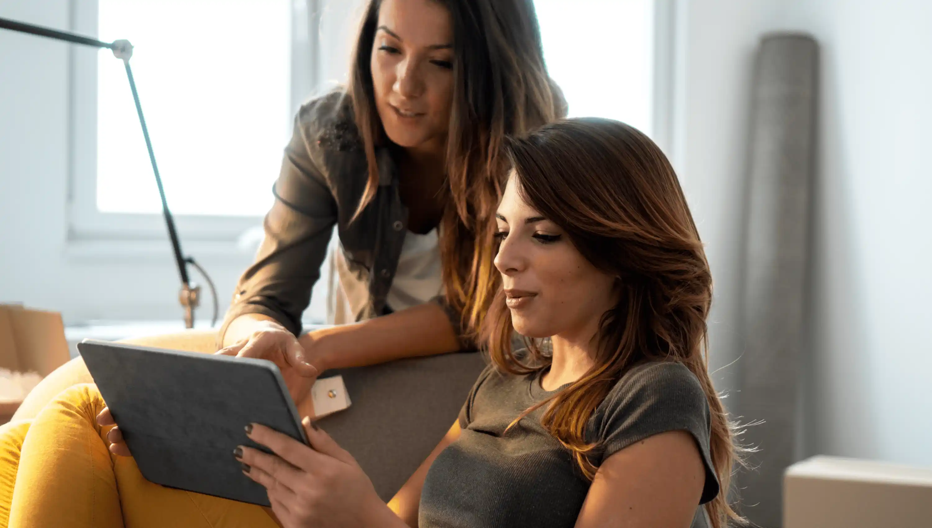 Two women with long hair looking at a tablet together in a bright room, one sitting on a couch and the other leaning over her shoulder.