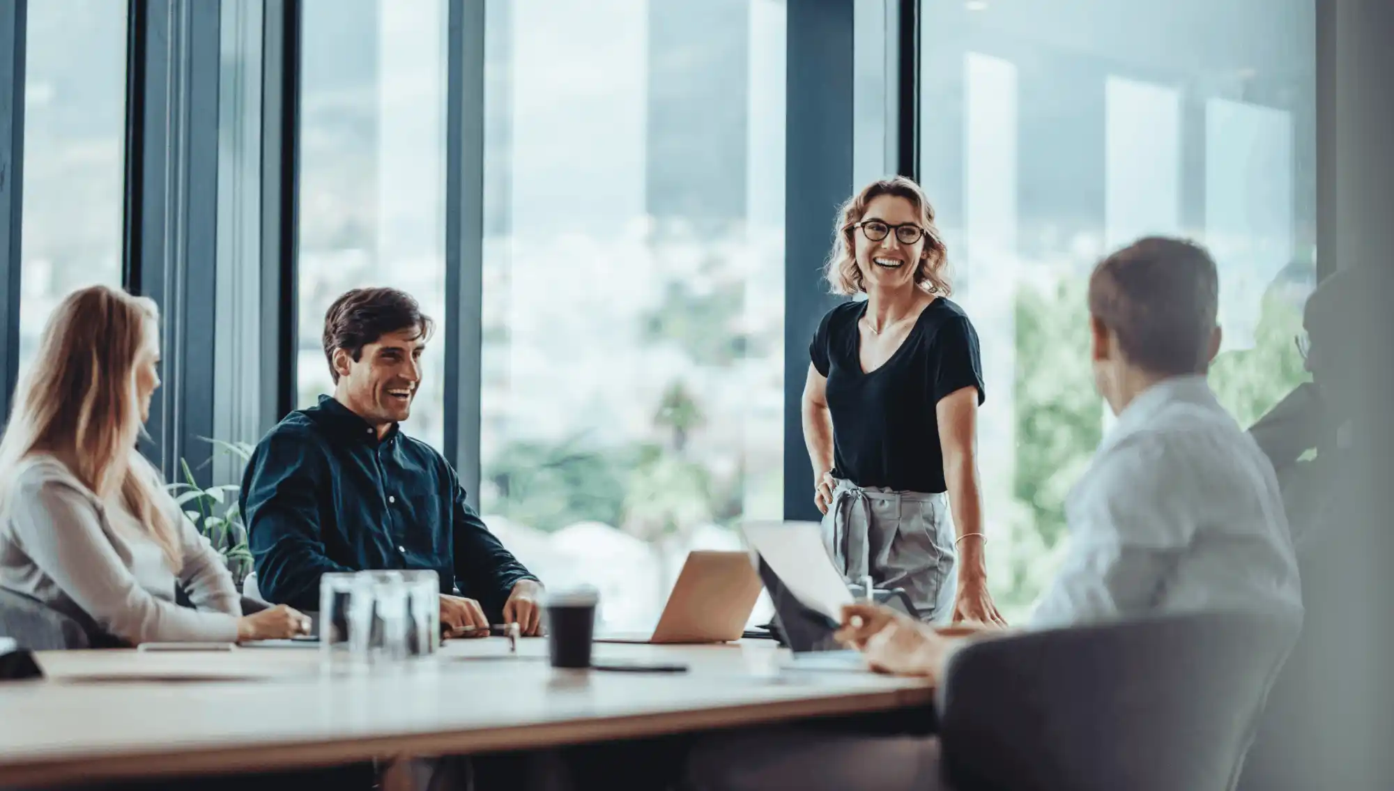 Four business professionals in a modern office with large windows, engaged in a lively meeting around a conference table, smiling and interacting with each other.