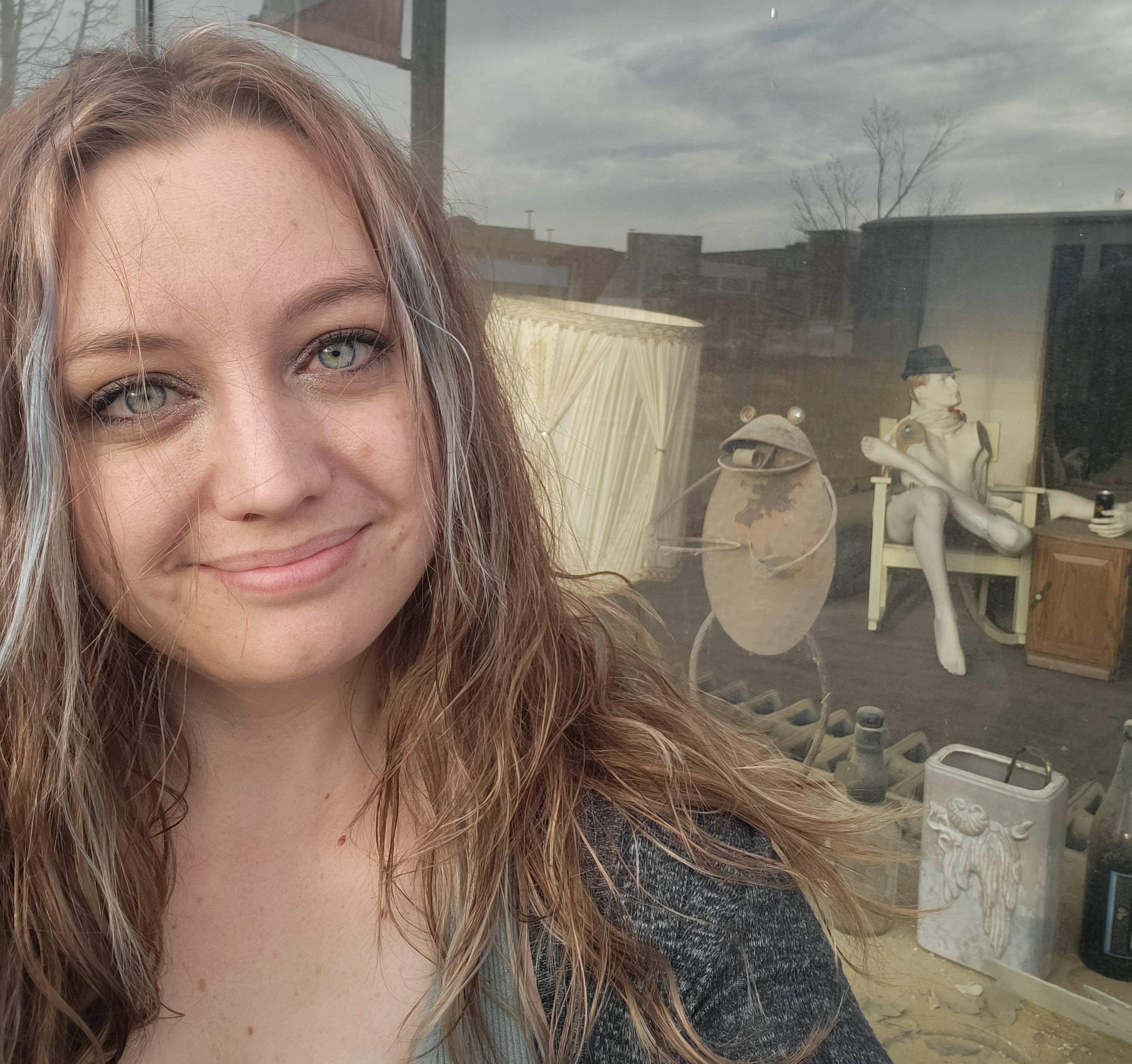 close up Image of Kassidi Andrus' face, standing in front of a vacant storefront with very strange items in the window i.e. a dancing metal frog, and a mannequin sitting in a chair wearing a fedora. 