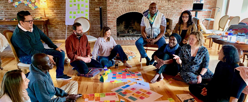 Group of diverse people sitting in a circle on the floor around colorful sticky notes and papers during a collaborative workshop session in a rustic brick-walled room.