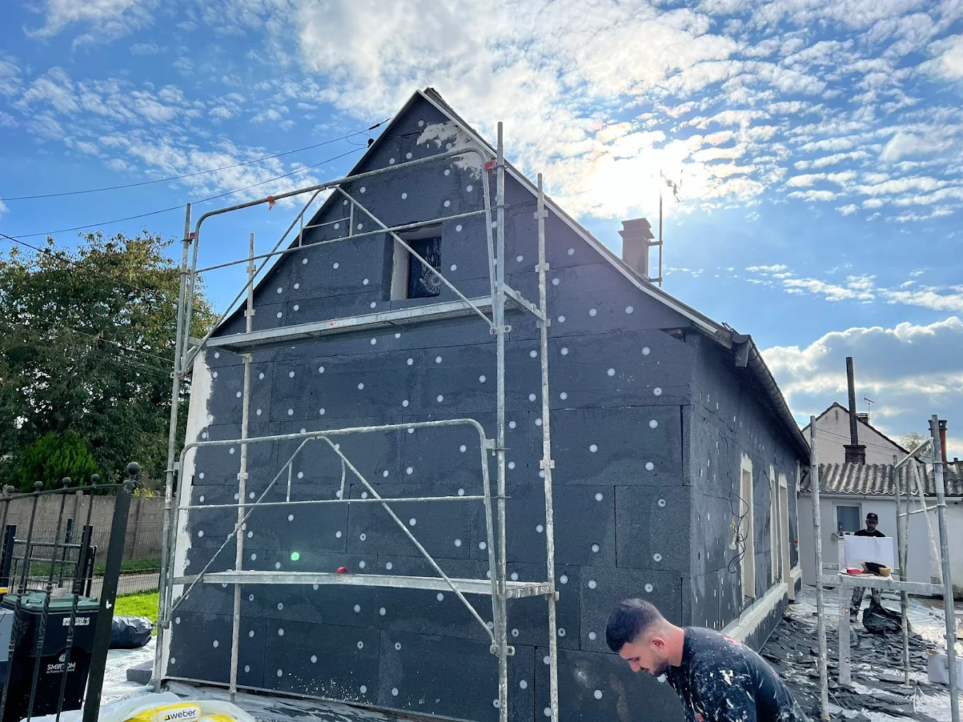 Vue aérienne d'une ville bordée par une rivière avec de nombreuses maisons, des arbres verts et un ciel partiellement nuageux.
