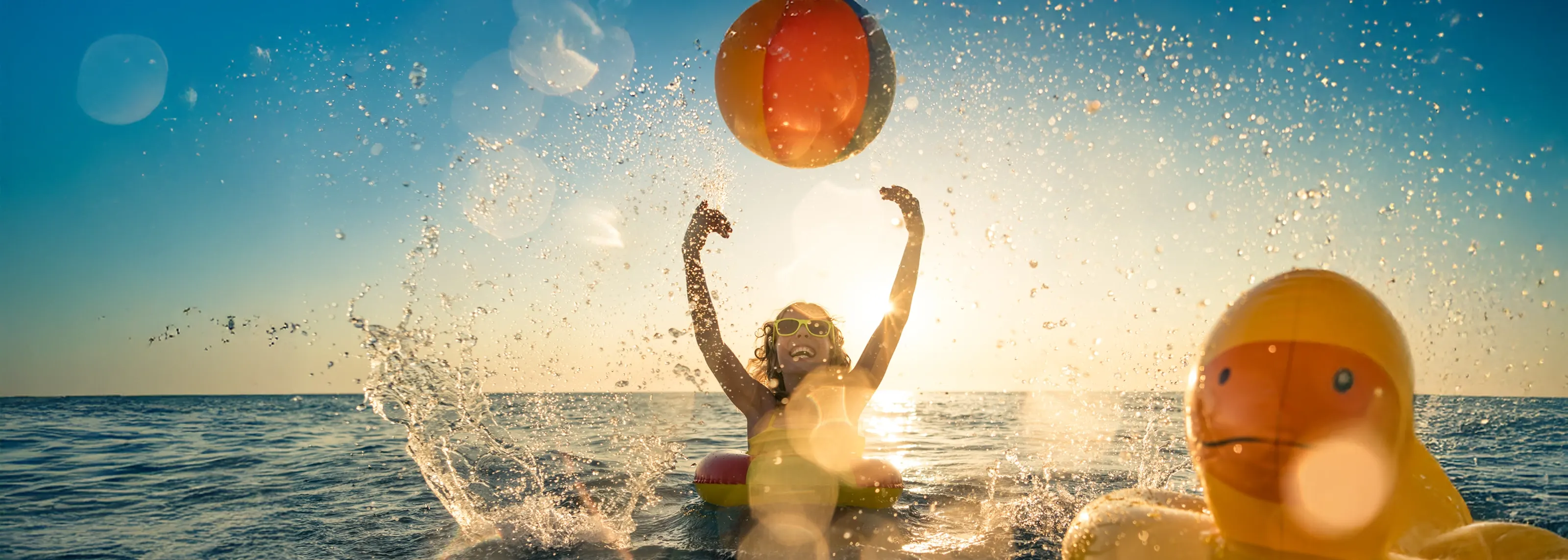 Child sitting in an inflatable ring in the sea, throwing a beach ball at sunset
