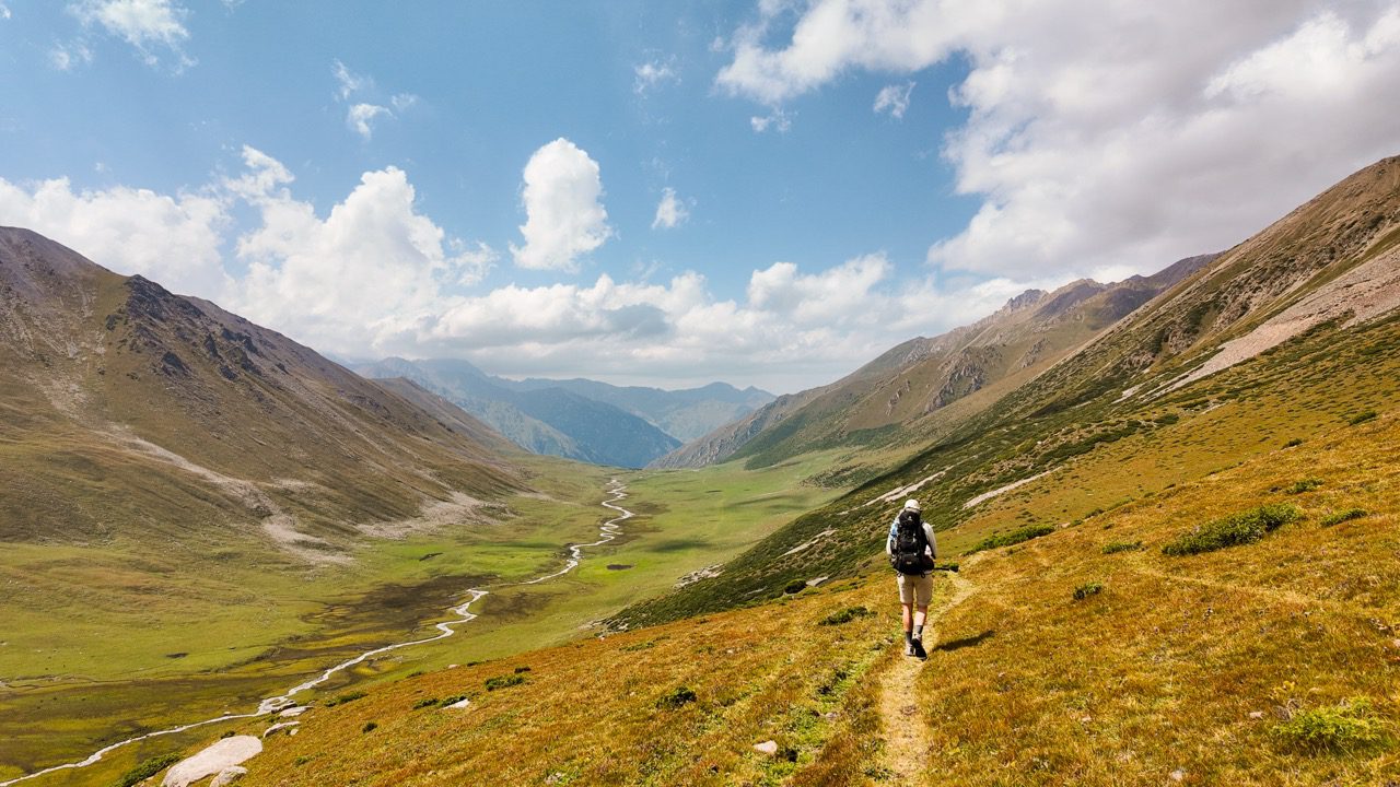 Hiker with a backpack walking a narrow trail high above a green mountain valley