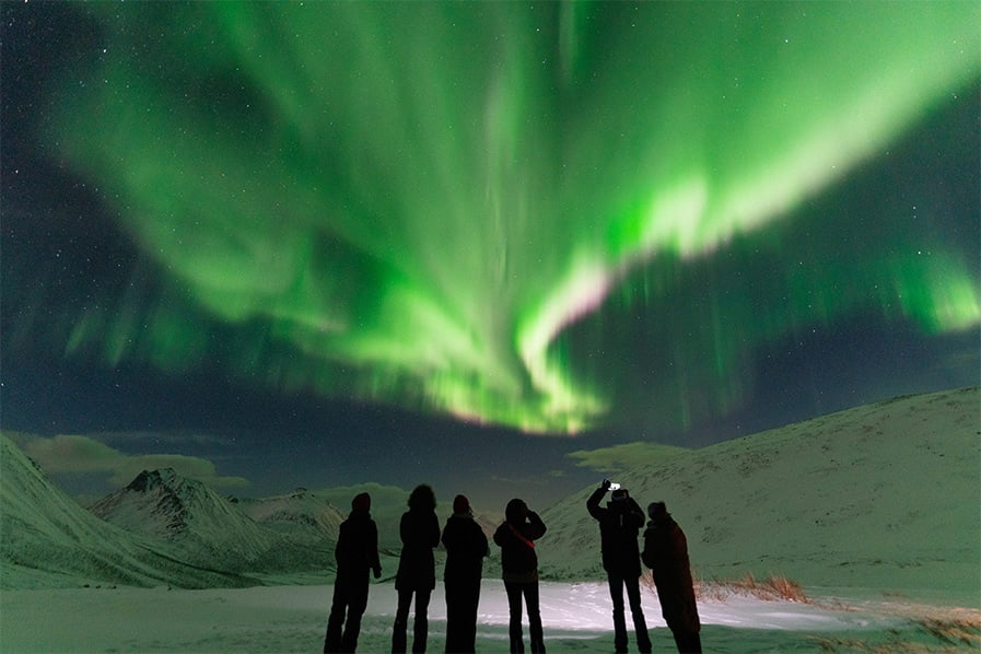 Small group of travellers watching green northern lights over snow-covered mountains