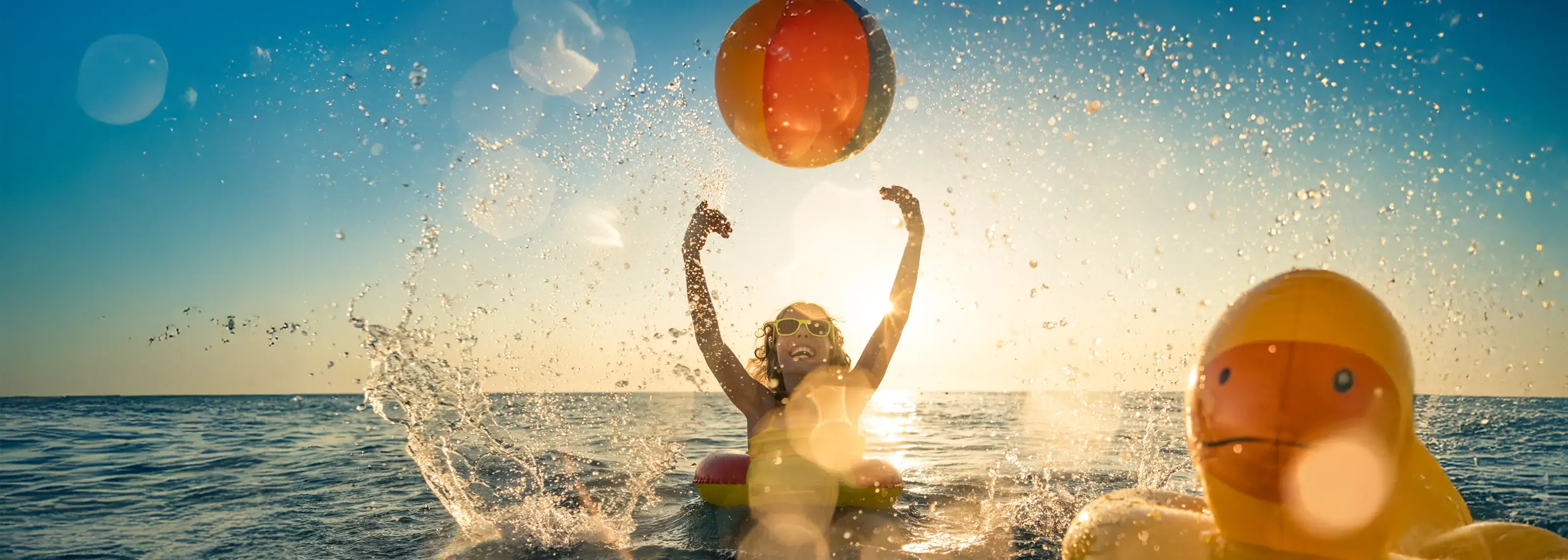 Child sitting in an inflatable ring in the sea, throwing a beach ball at sunset