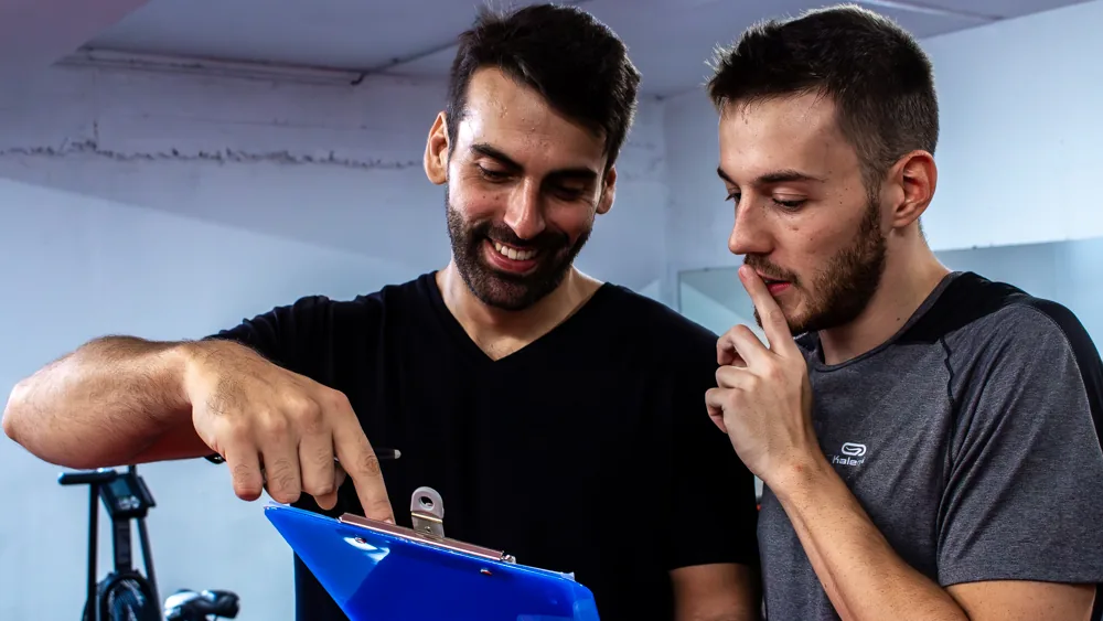 Nicolas, coach, montre une feuille sur un porte-bloc à un élève pensif, en salle de sport.