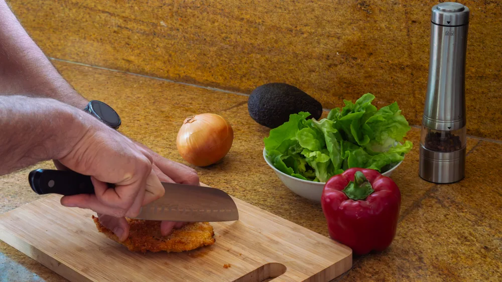 Des mains coupent un aliment sur une planche en bois, entourées d'oignon, d'avocat, de salade et de poivron rouge.