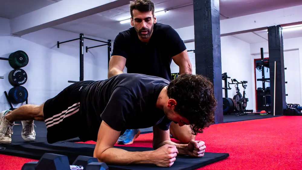 Un élève en gainage sur un tapis, Nicolas debout derrière lui le surveille, dans une salle de sport.