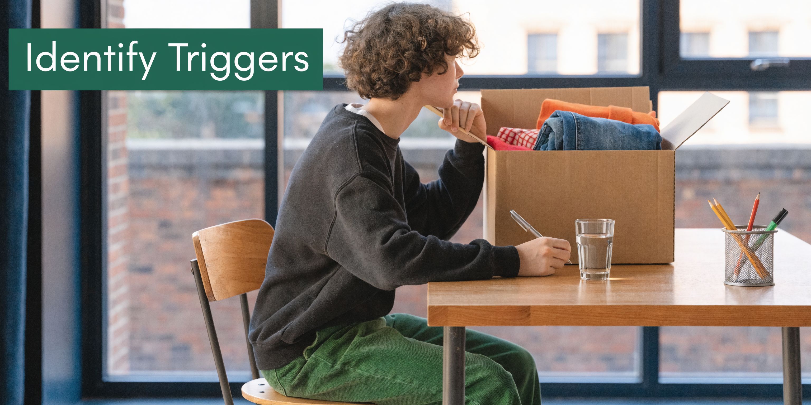 A person sitting at a desk thoughtfully writing in a notebook next to a cardboard moving box.