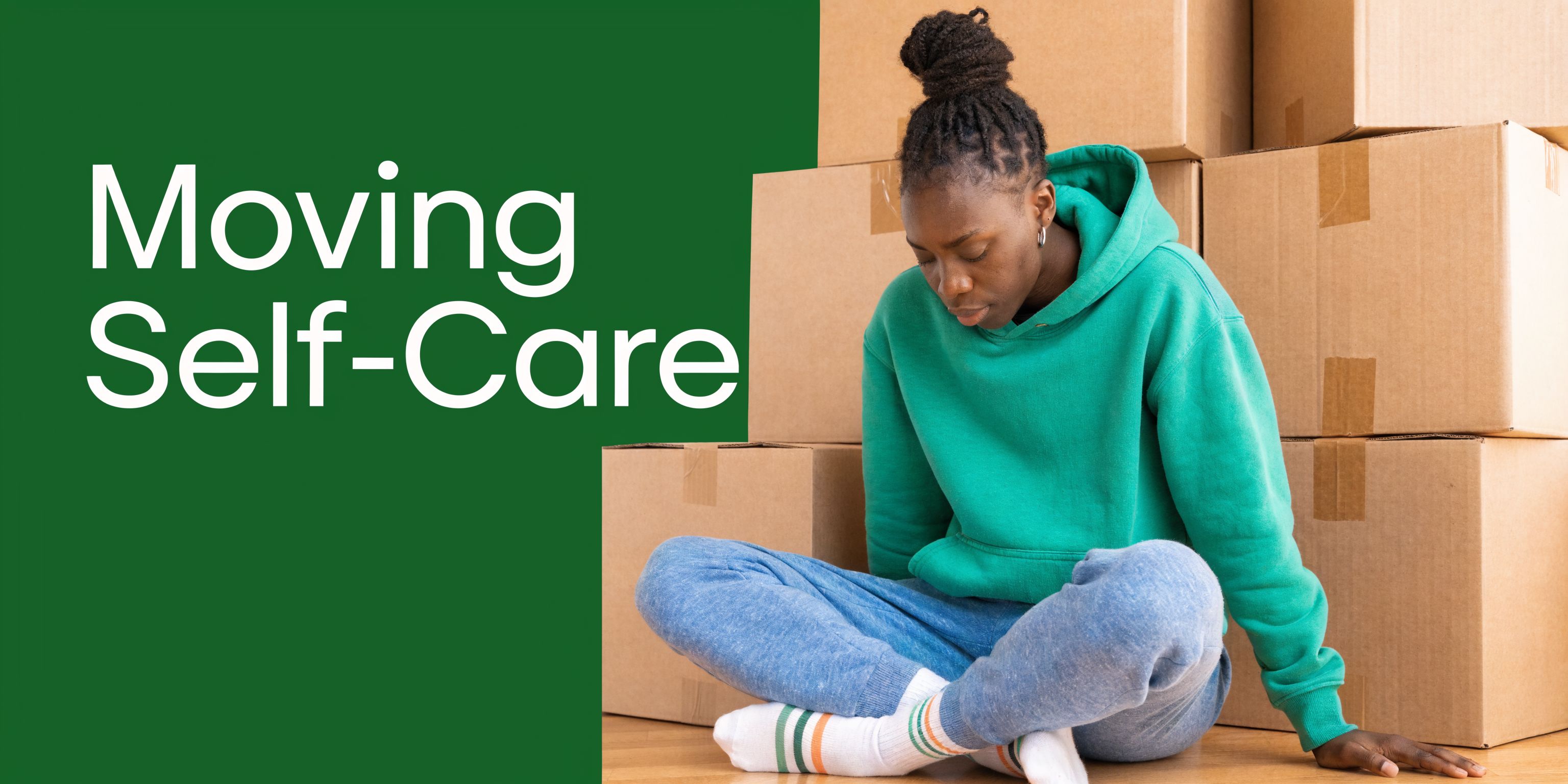 A woman with her head down sits on the floor surrounded by stacked cardboard moving boxes.