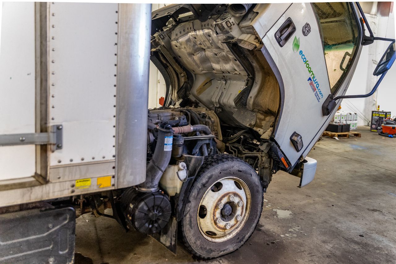 Semi-truck in a repair shop with cab tilted forward, exposing engine components for maintenance, showing wheel and surrounding workshop area. 