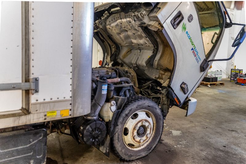 Semi-truck in a repair shop with cab tilted forward, exposing engine components for maintenance, showing wheel and surrounding workshop area. 