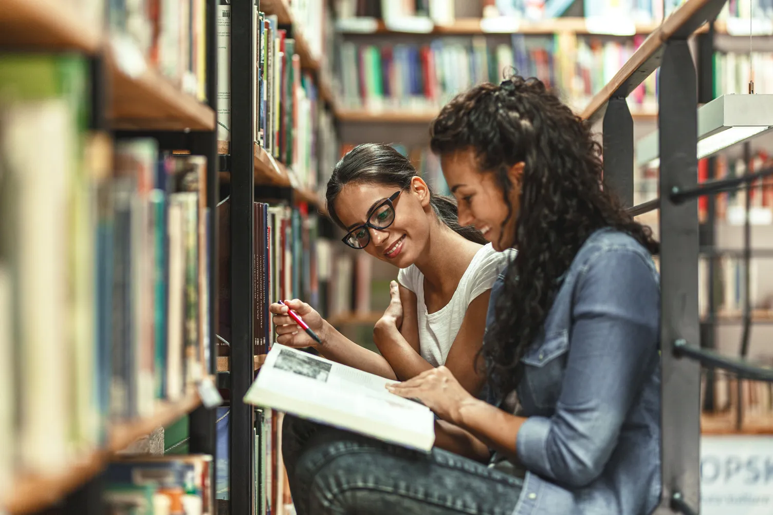 Two female students sitting on the floors of a library as they look at a book together.