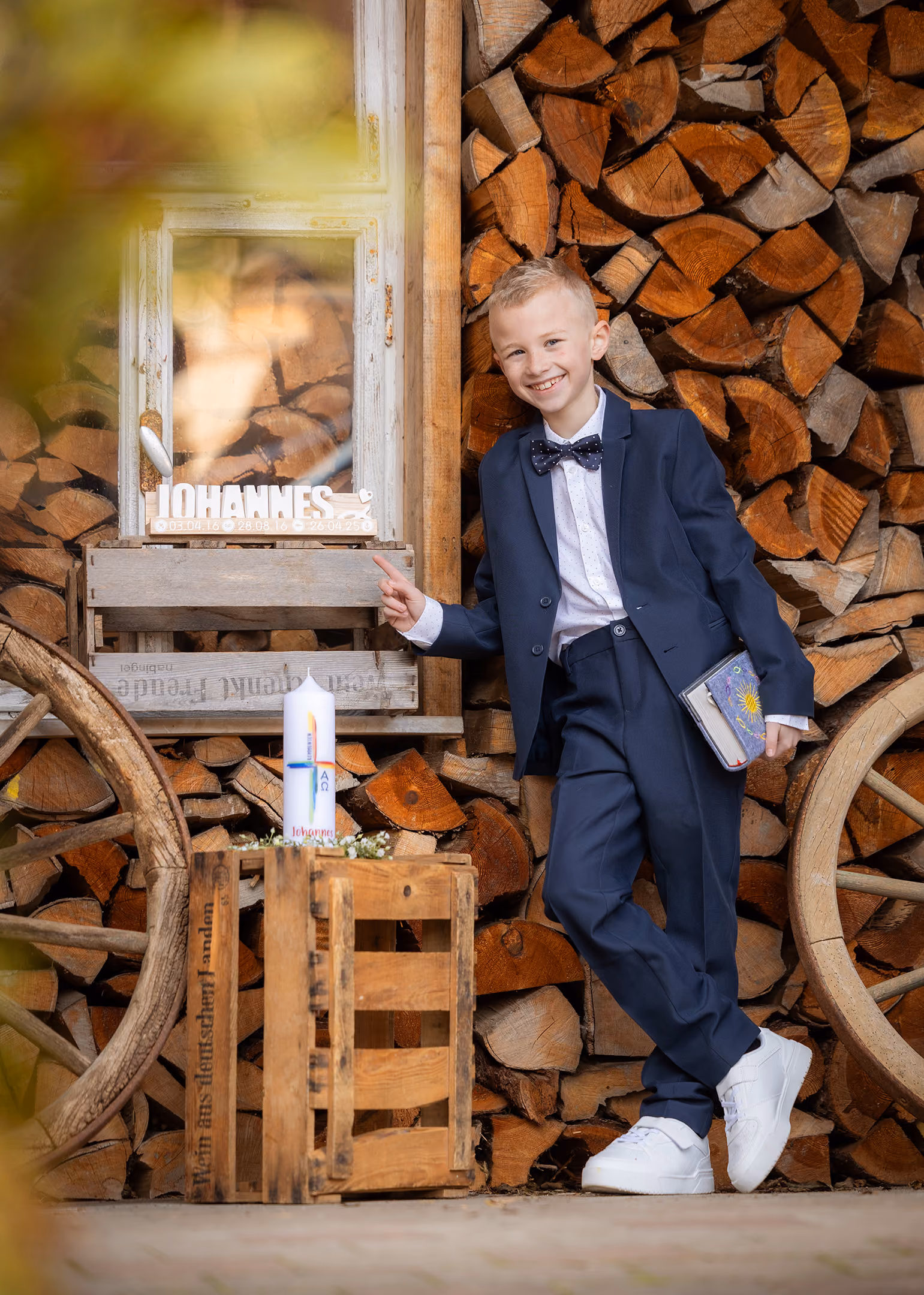 Lächelnder Junge im Anzug lehnt vor Kaminholz mit Kerze und Buch, zeigt auf Fenster mit Name Johannes.