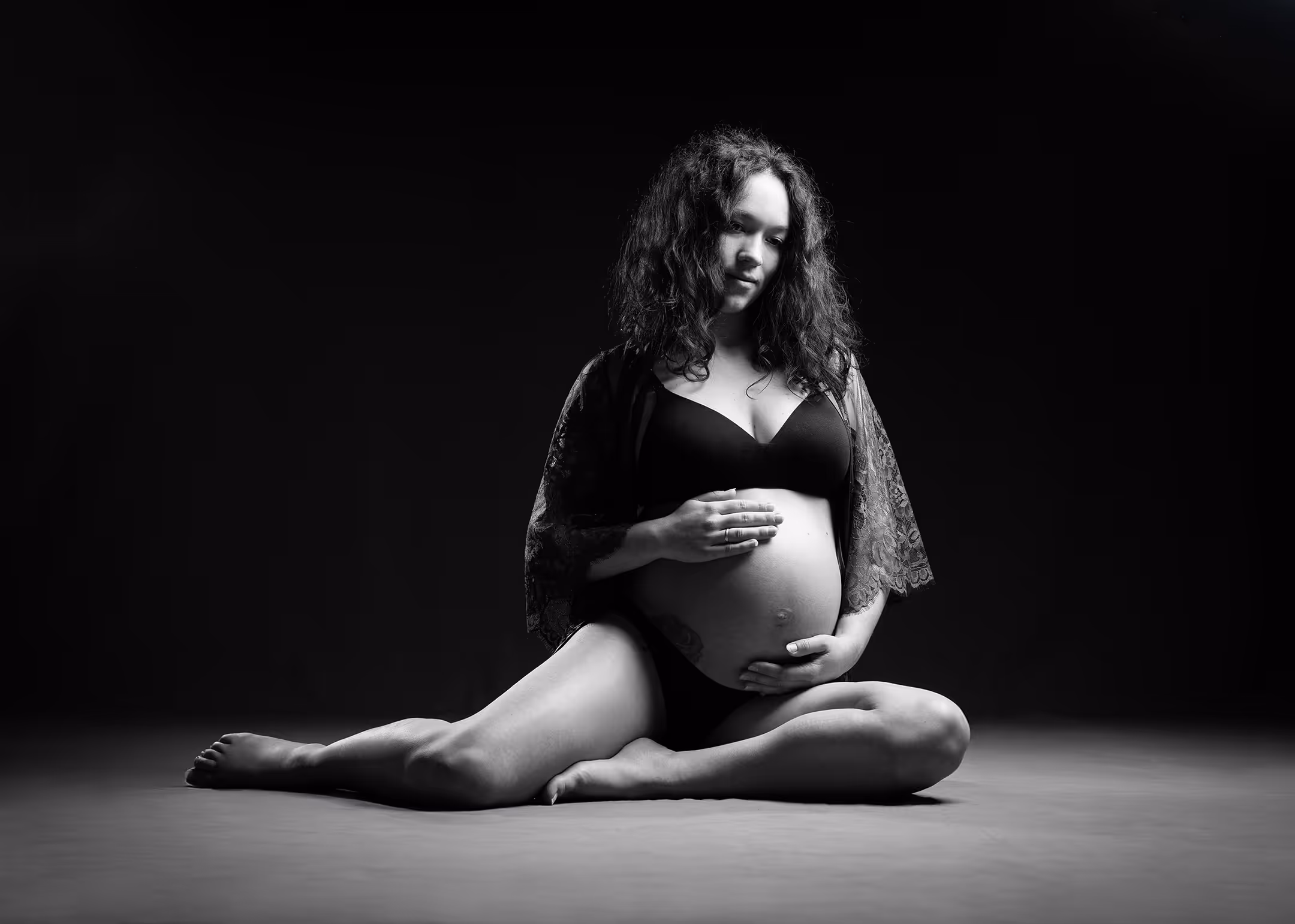 Schwangere Frau sitzt auf dem Boden, hält sanft ihren Bauch, umgeben von dunklem Hintergrund.