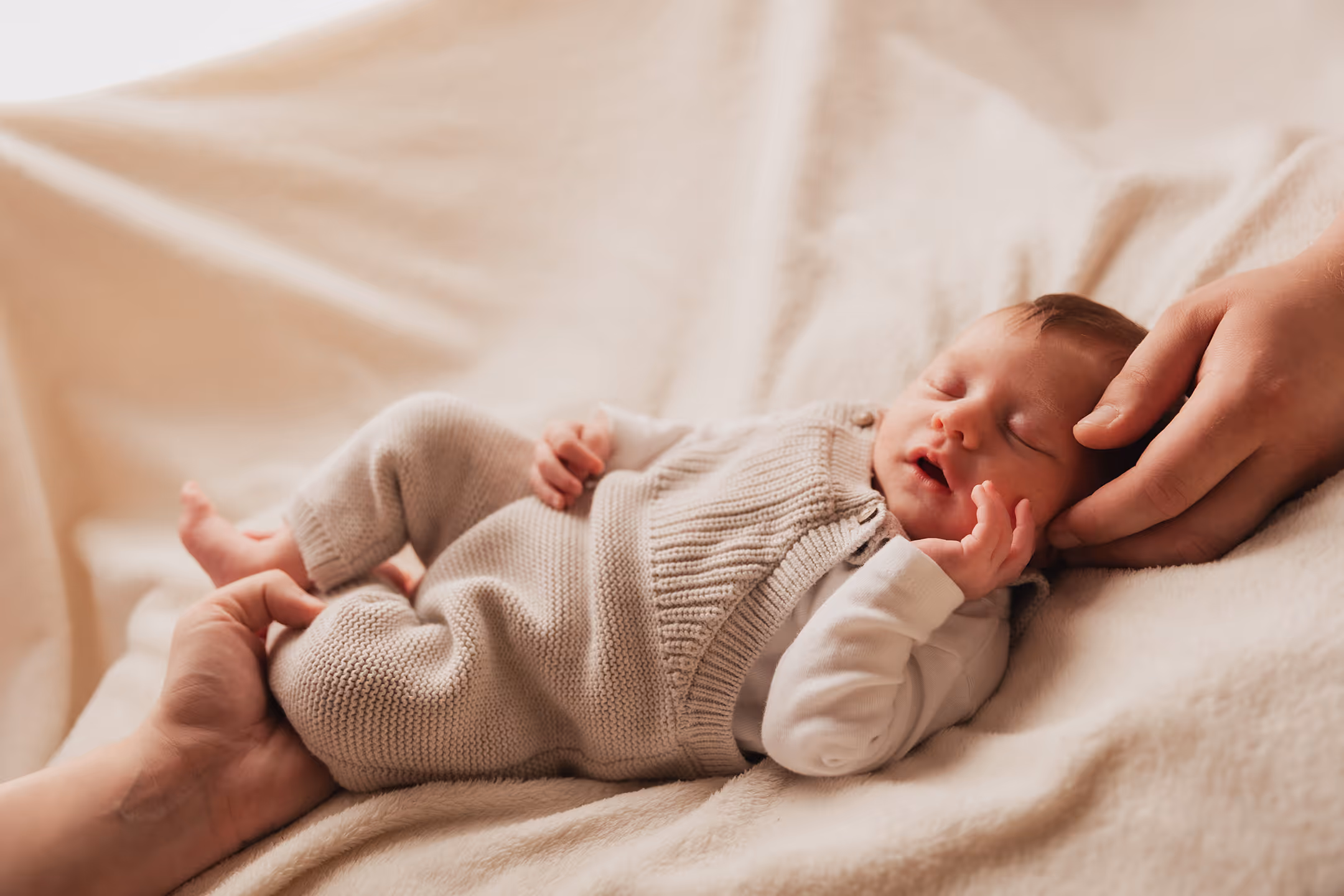 Neugeborenes Baby liegt schlafend auf einer weichen Decke, wird sanft von zwei Händen gehalten.
