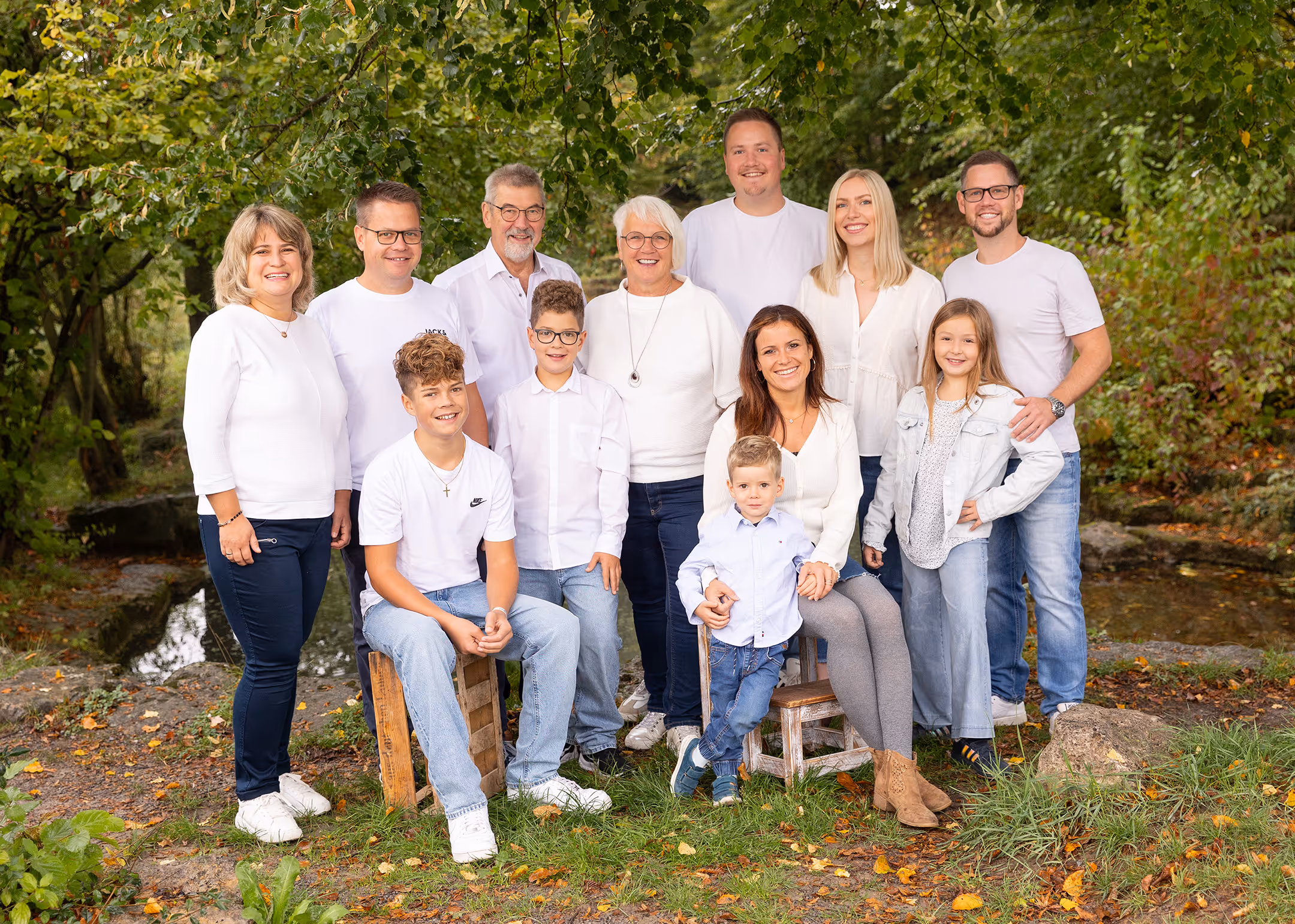 Große Familie mit acht Erwachsenen, drei Kindern und einem Kleinkind, alle lächelnd und teils sitzend, teils stehend in einem grünen Park mit Herbstlaub.