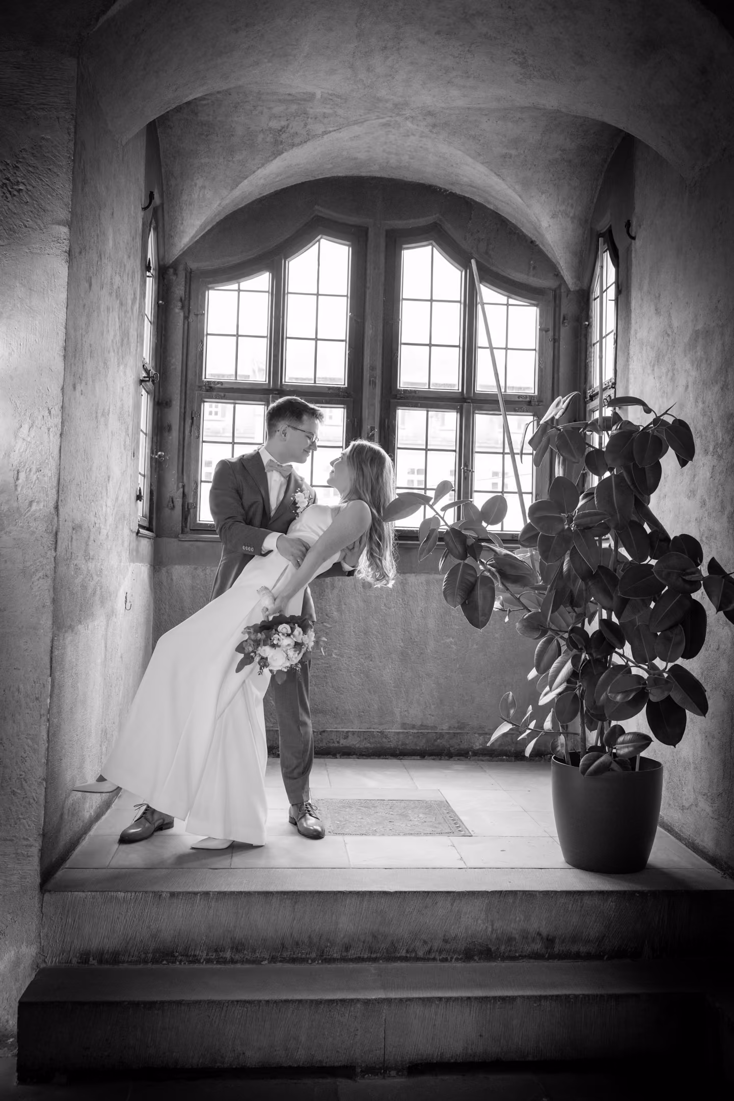 Hochzeitspaar in einem historischen Raum mit Bogenfenster, Braut lehnt sich zurück, hält Blumen.
