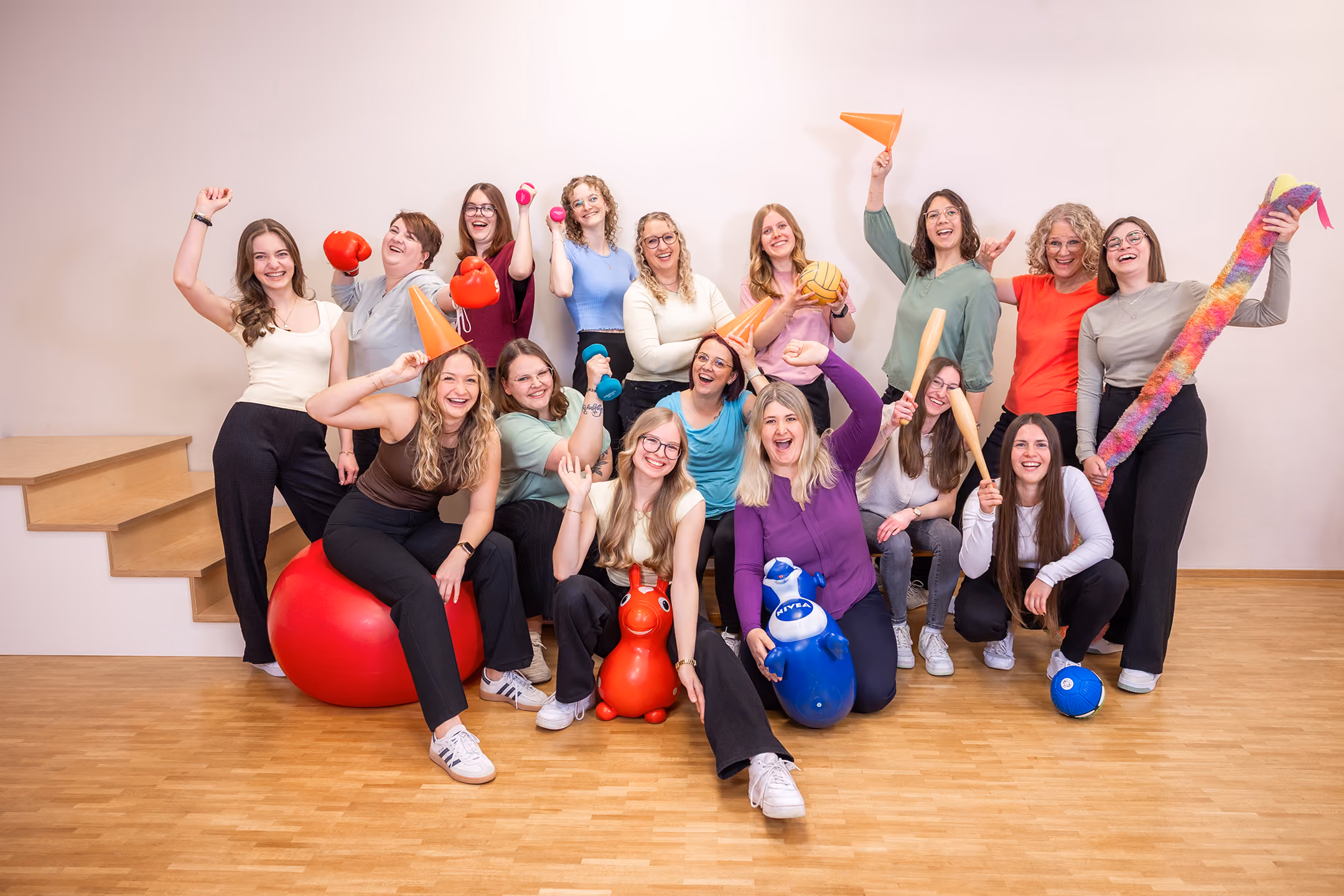 Gruppenfoto von 16 fröhlichen Frauen in lässiger Kleidung mit Sport- und Spielutensilien im weißen Raum.