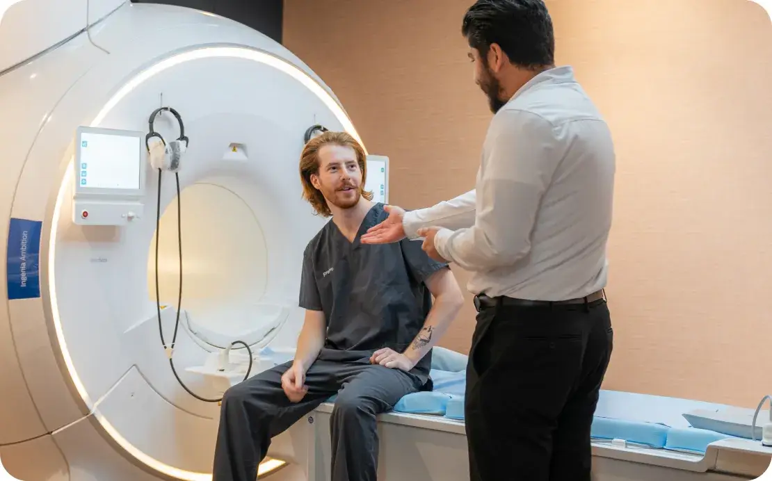 A man sitting on an MRI bed speaking with a technician
