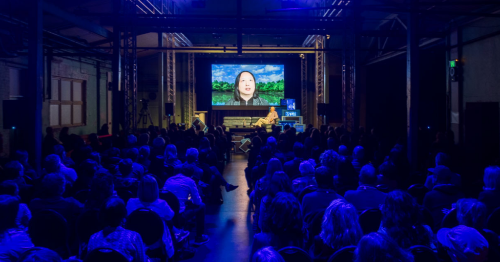 A room of people watching a panel, with one person on stage and one person on the screen