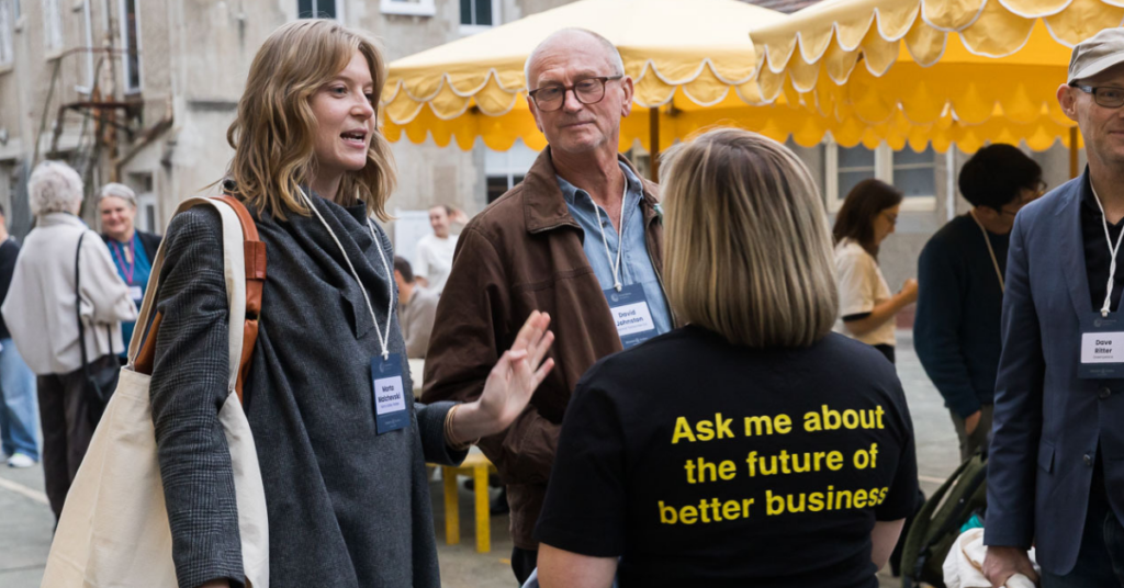 A woman wearing a t shirt that says “Ask me about the future of better business” talking to two people in a courtyard