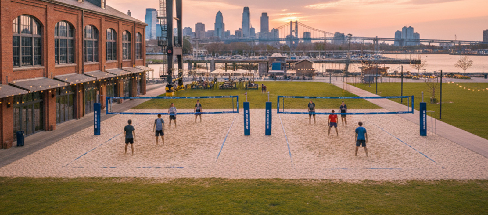 Two beach volleyball courts with players, old brick building on the left, city skyline and bridge in the background at sunset.