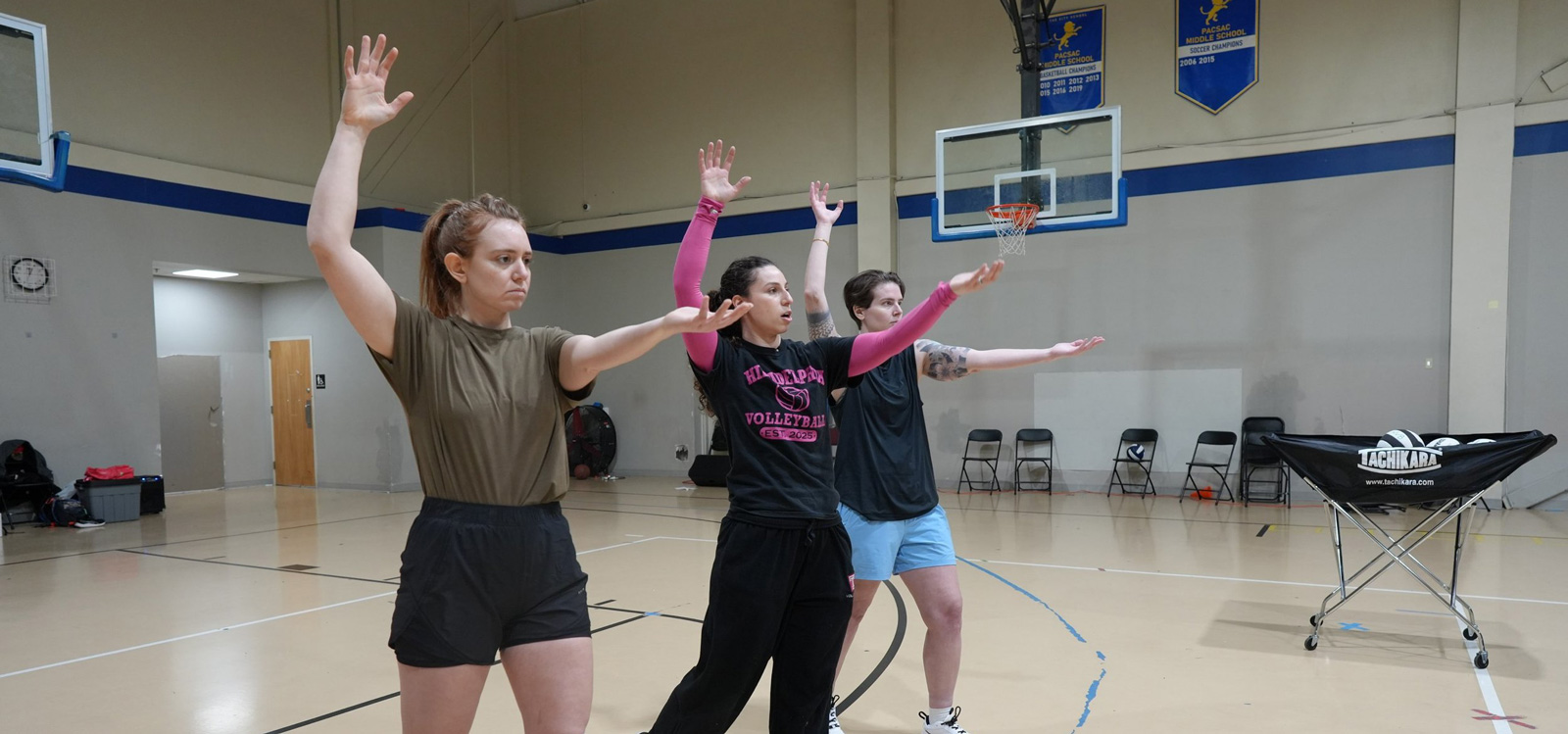 Three women standing in a gymnasium with arms raised, practicing volleyball drill near a basketball hoop and a cart of volleyballs.