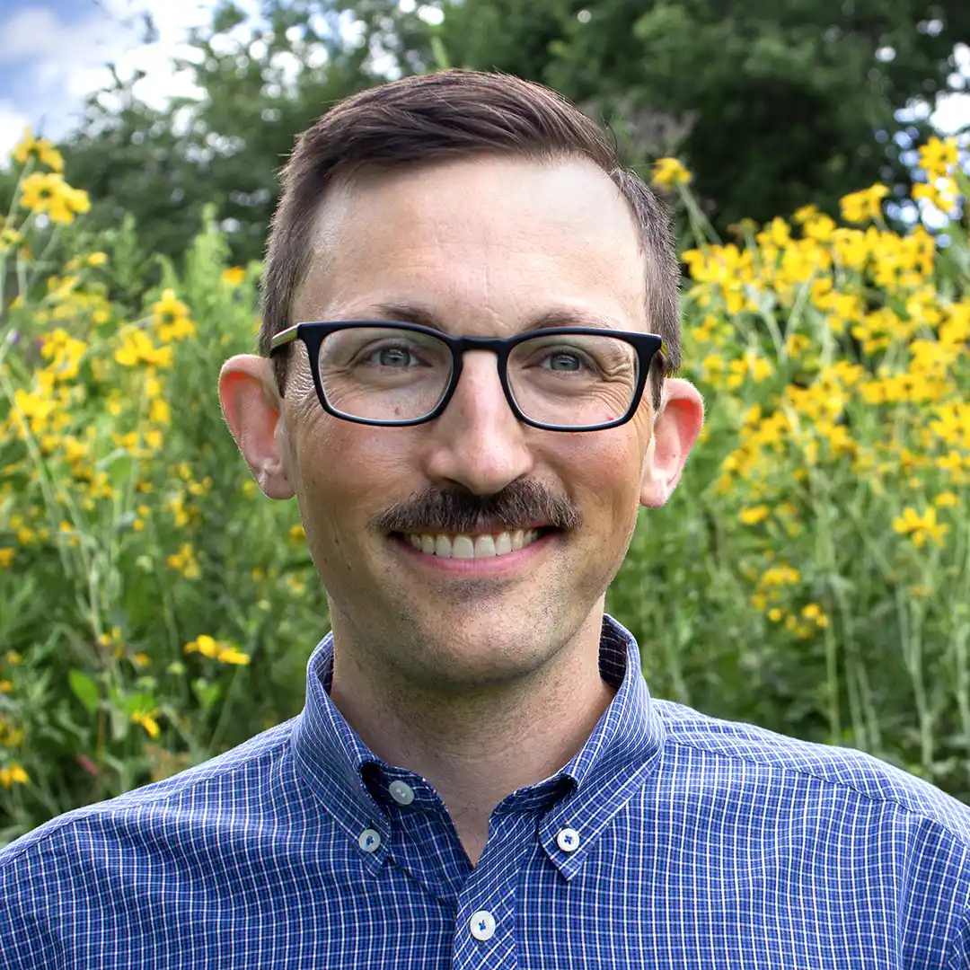 A portrait photo shot outside of Danny Harms against a glass store window wearing black rim glasses, a hat with a treeline graphic, white collared shirt, and black zip-up hoodie