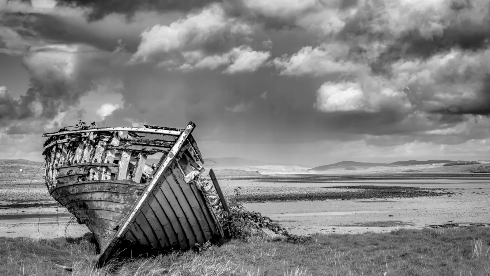 Black-and-white photo of a weathered boat on a quiet shore beneath cloudy skies.