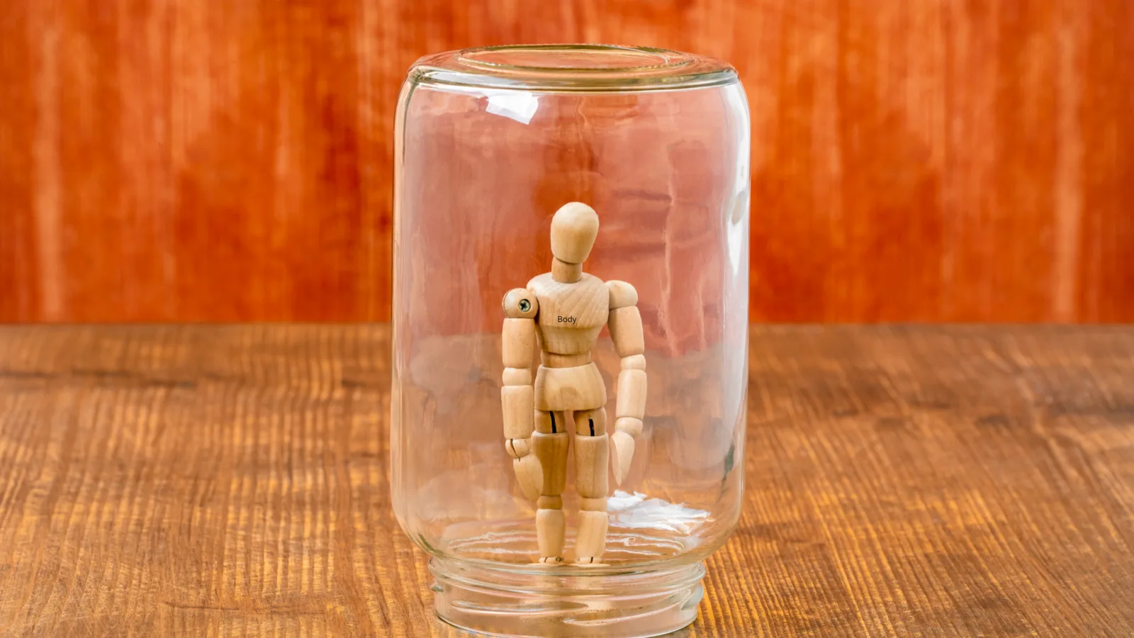 Wooden figure standing inside a glass jar on a wooden table.