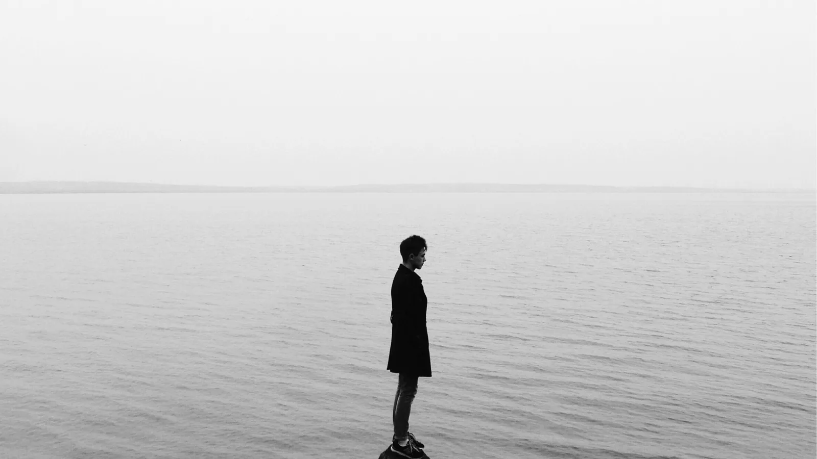 Person in a dark coat standing alone on rocks beside calm water.