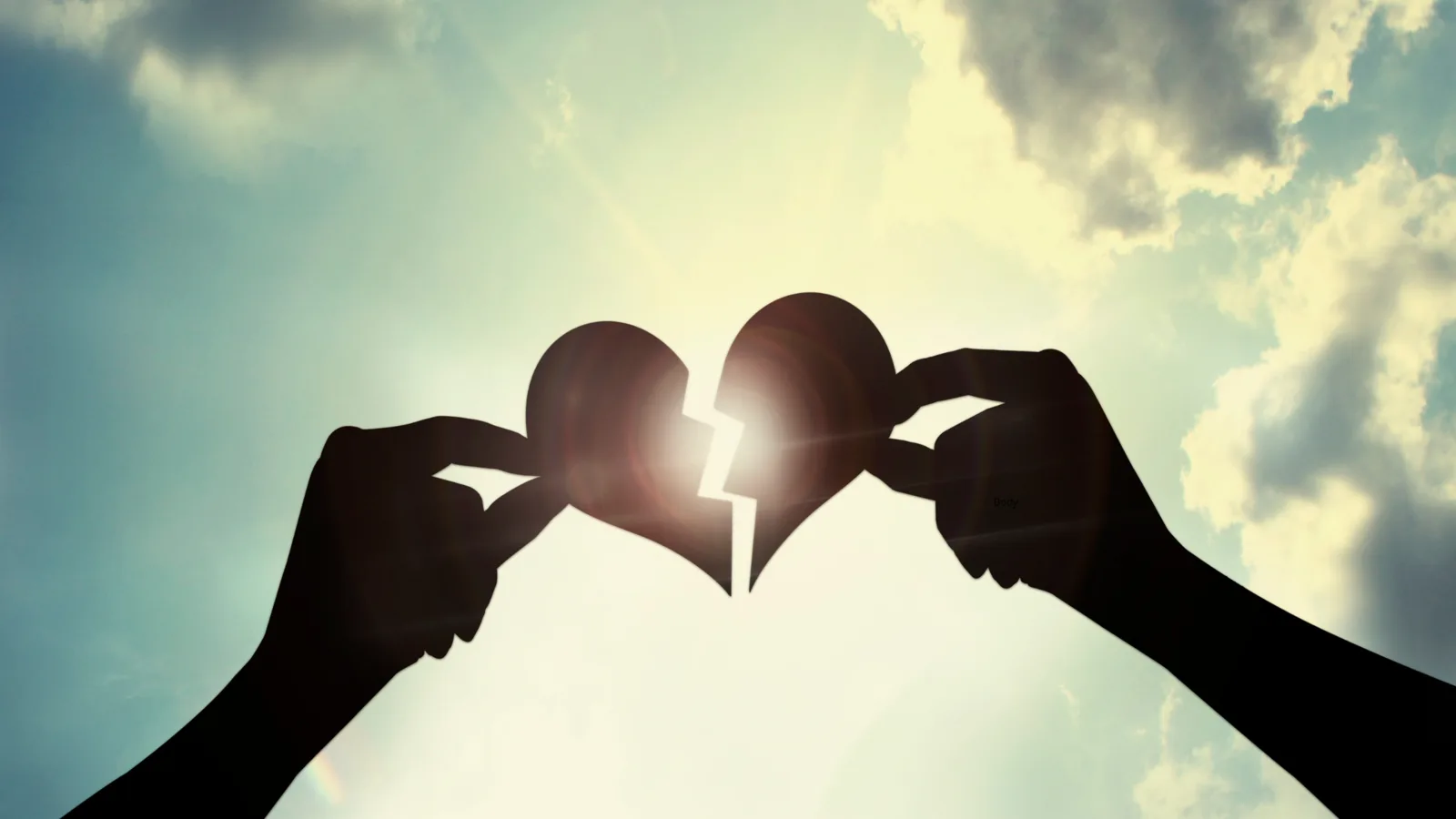 Two hands holding halves of a broken heart against the sky.