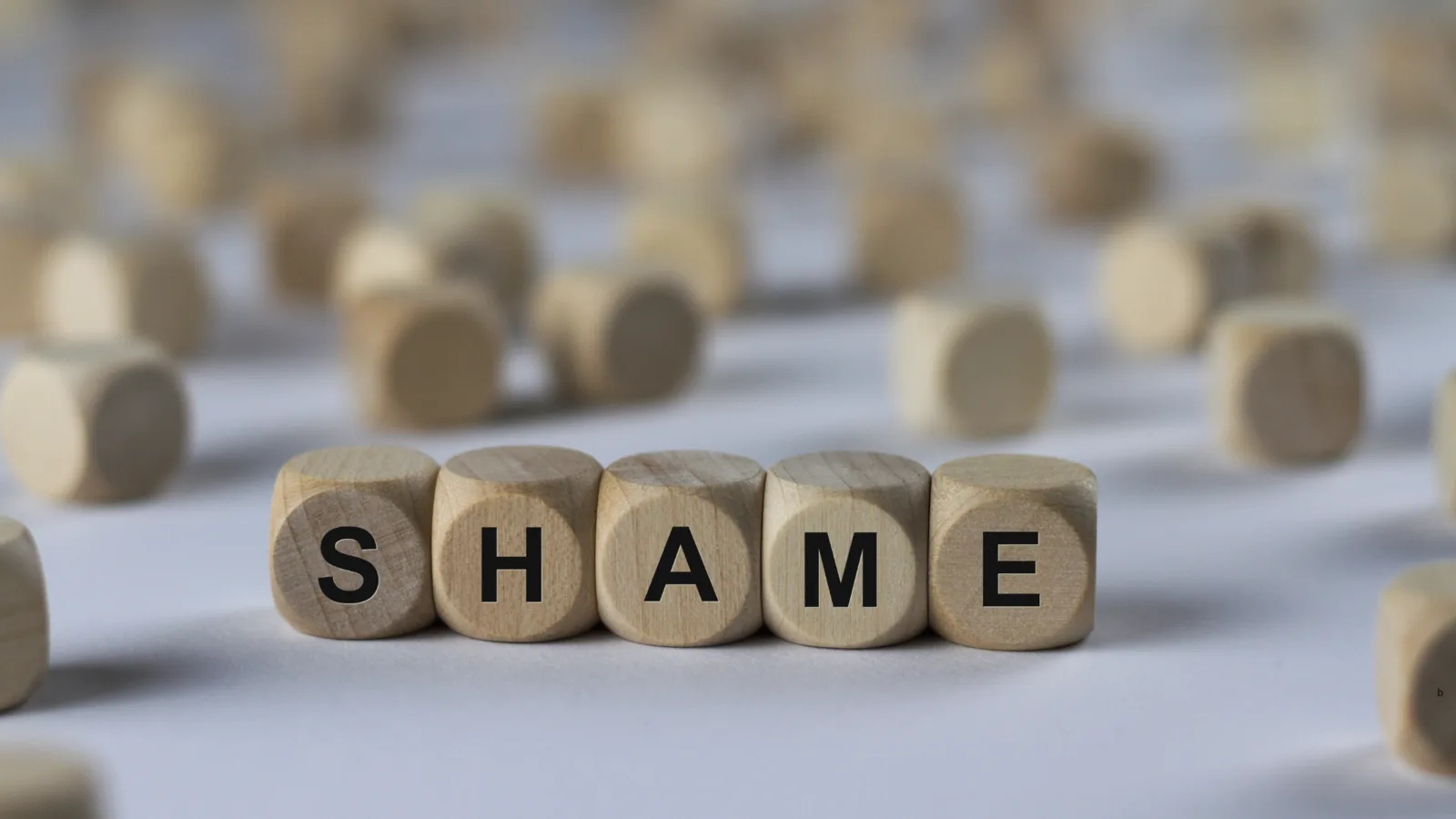 Wooden letter blocks spelling “shame” on a softly blurred surface.