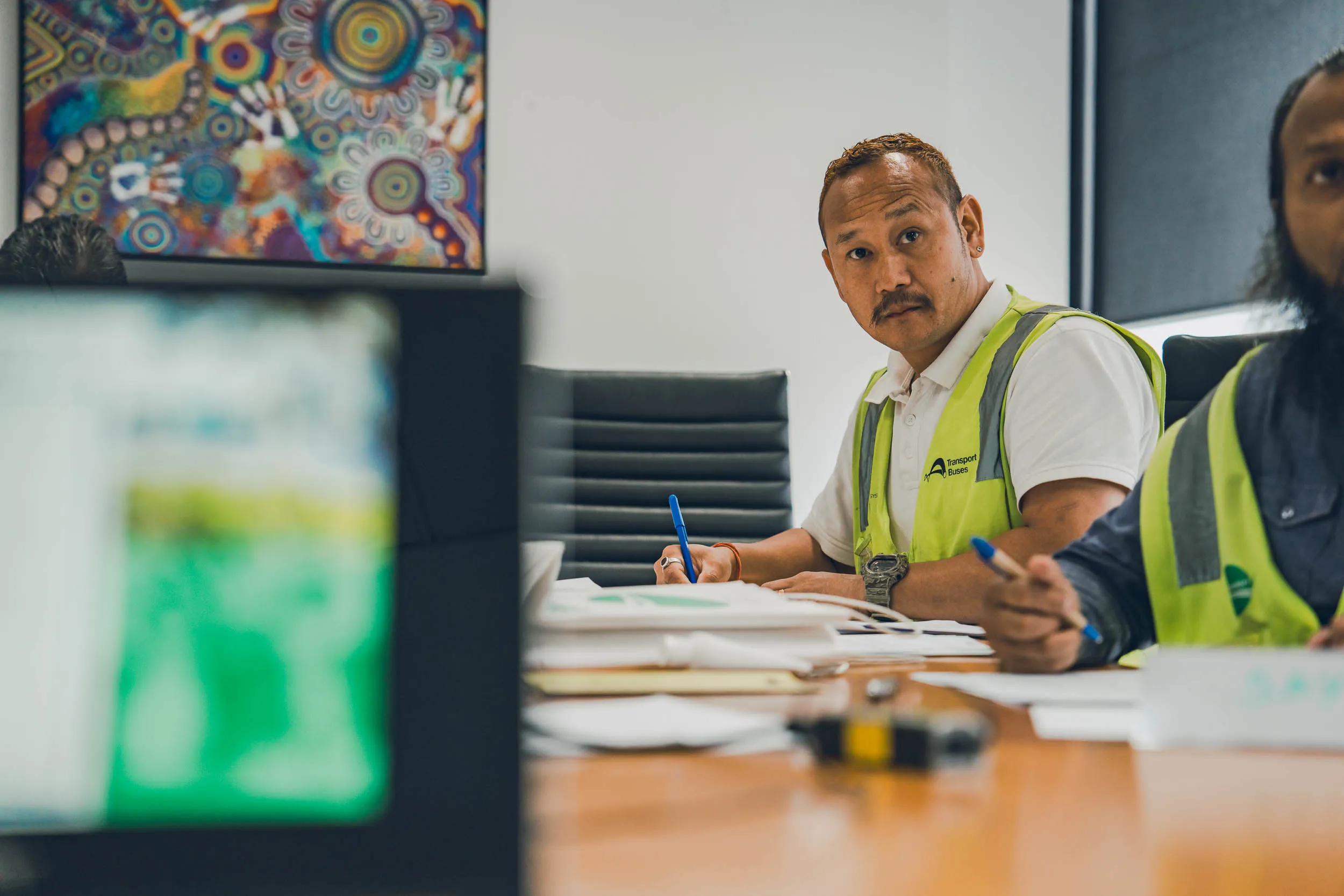 Man in a high-visibility vest writing with a pen at a meeting table with papers and another man.
